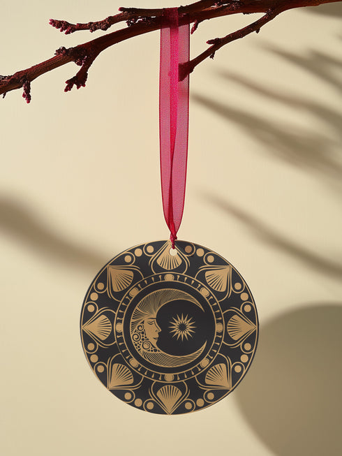 Round metal ornament featuring a golden crescent moon face with star detail, surrounded by art deco shell motifs on black background. 3.5-inch diameter double-sided design with lunar phase pattern and crimson ribbon attachment. Vintage-inspired celestial artwork.
